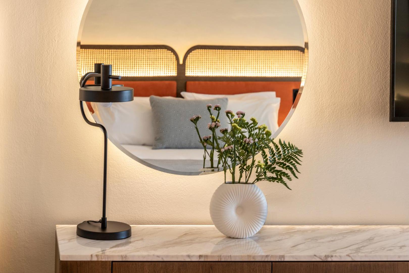 Kerkyra Blue Hotel & Spa numura interjers ar apaļu spoguli, kurā atspoguļojas gulta ar oranžu galvgali un dekoratīviem ziediem priekšplānā.