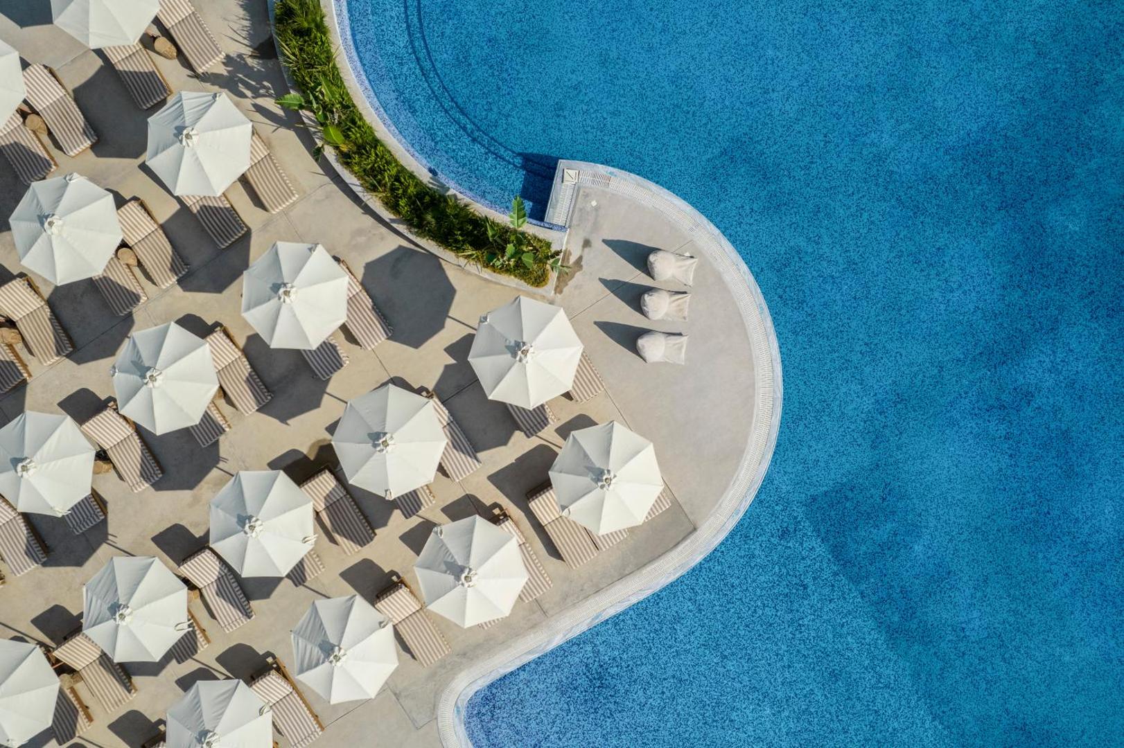 Kerkyra Blue Hotel & Spa skats no augšas uz plašu zilu baseinu un sakārtotām sauļošanās zonām ar gaišiem saulessargiem un zviļņiem.