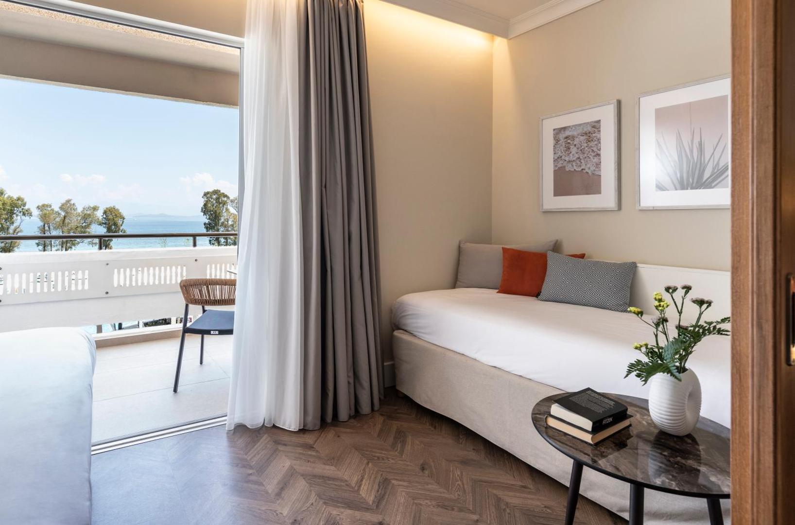 Kerkyra Blue Hotel & Spa gaišs numurs ar balkonu, no kura paveras skats uz Jonijas jūru un zaļojošiem kokiem Korfu piekrastē.