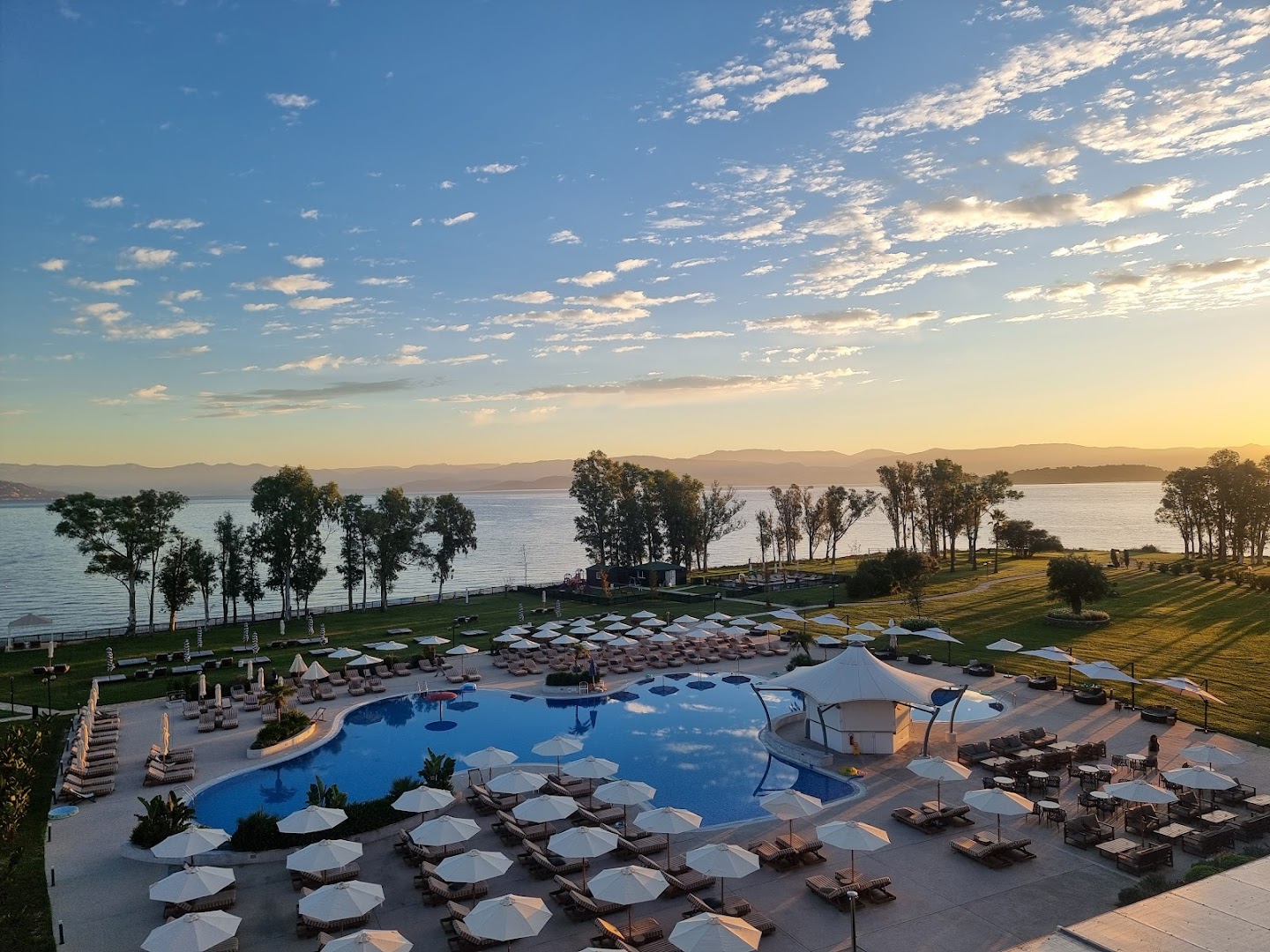 Panorāmas skats uz Kerkyra Blue Hotel & Spa baseinu un dārzu saulrietā ar skatu uz jūru un kalniem tālumā