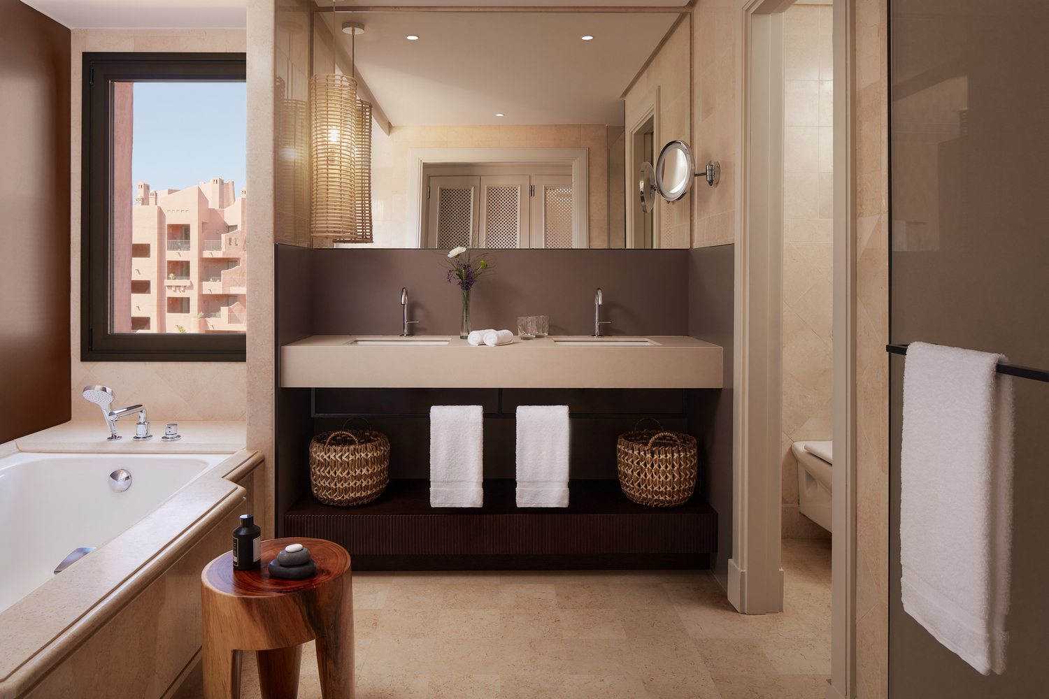 Tivoli La Caleta Tenerife Resort luksusa numura vannas istaba ar divām izlietnēm, vannu un skatu pa logu uz kūrorta arhitektūru