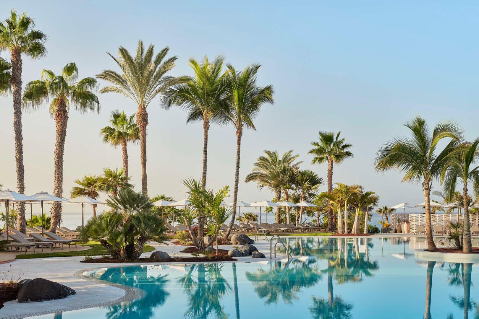 Tivoli La Caleta Tenerife Resort baseina skats uz okeāna pusi ar augstām palmām un baltiem saulessargiem rīta gaismā.
