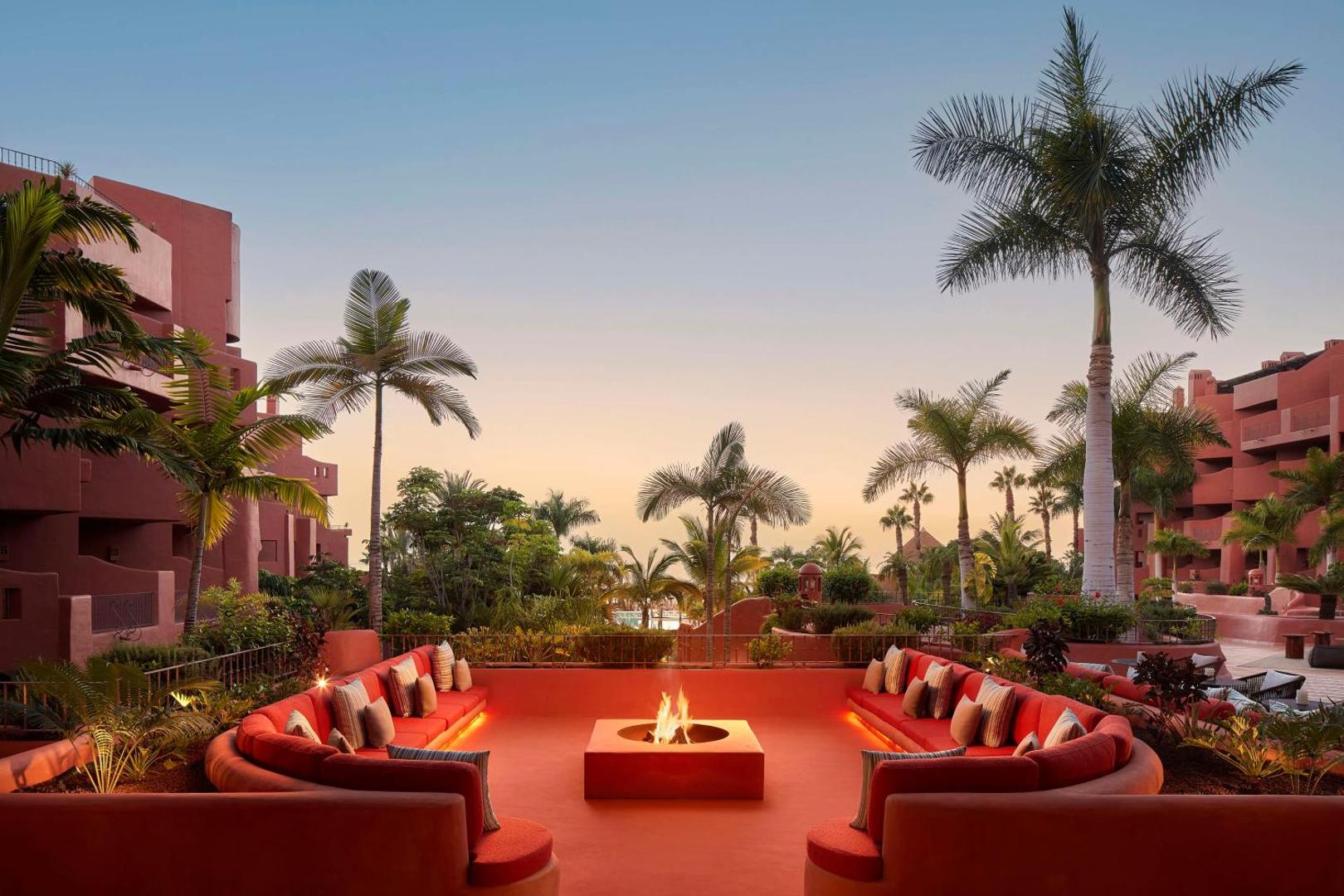 Tivoli La Caleta Tenerife Resort āra atpūtas zona ar ugunskura vietu, sarkaniem dīvāniem un palmām saulrieta laikā