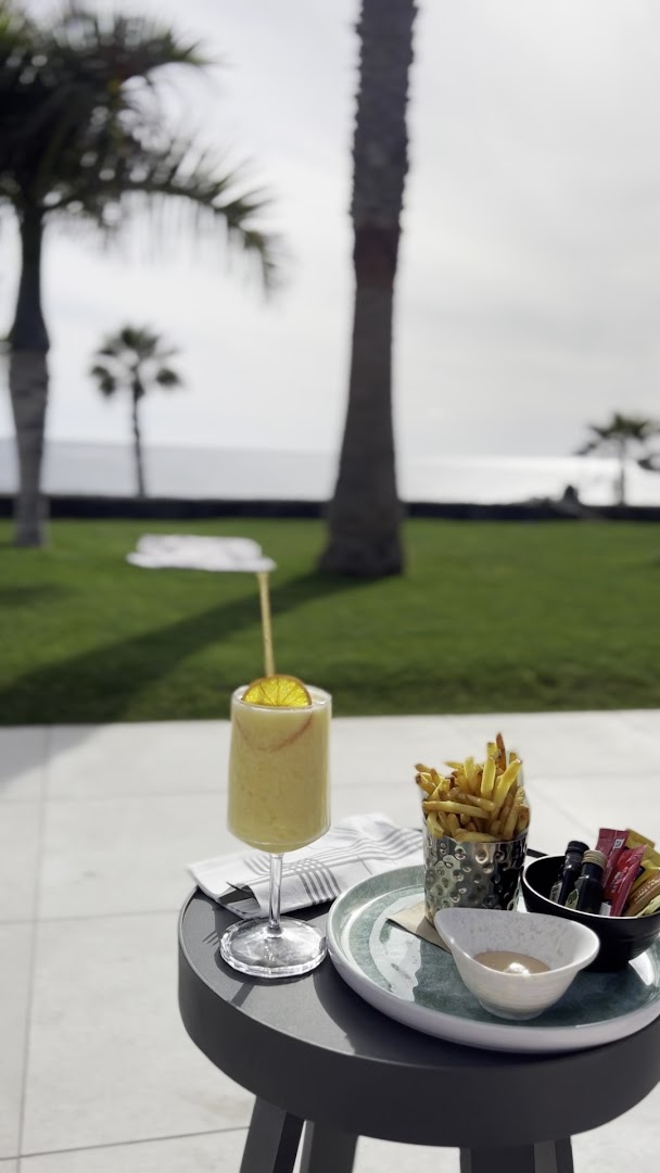Atsvaidzinošs kokteilis un uzkodas uz galdiņa Tivoli La Caleta Tenerife Resort āra terasē ar skatu uz palmu dārzu un okeānu