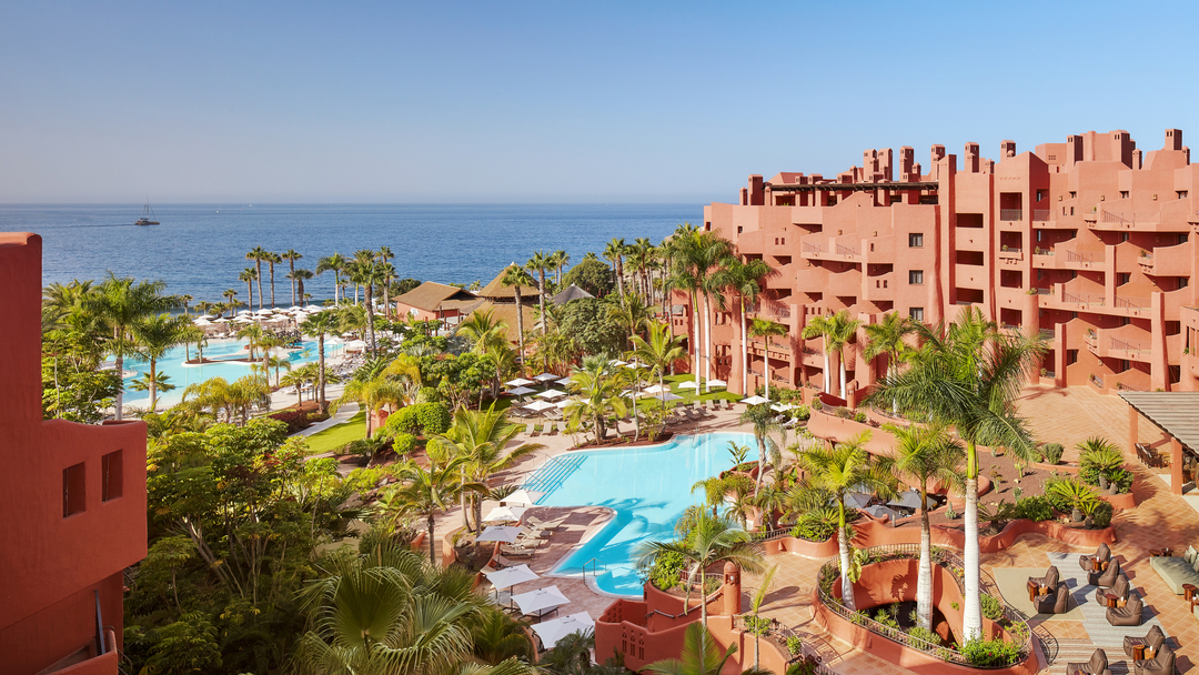 Tivoli La Caleta Tenerife Resort skats no augšas uz āra baseiniem, palmu dārzu un okeāna piekrasti saulainā dienā Tenerifē