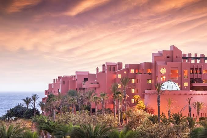 Tivoli La Caleta Tenerife Resort ēkas fasāde saulrieta gaismā ar palmām un okeānu fonā Tenerifē, Spānijā.