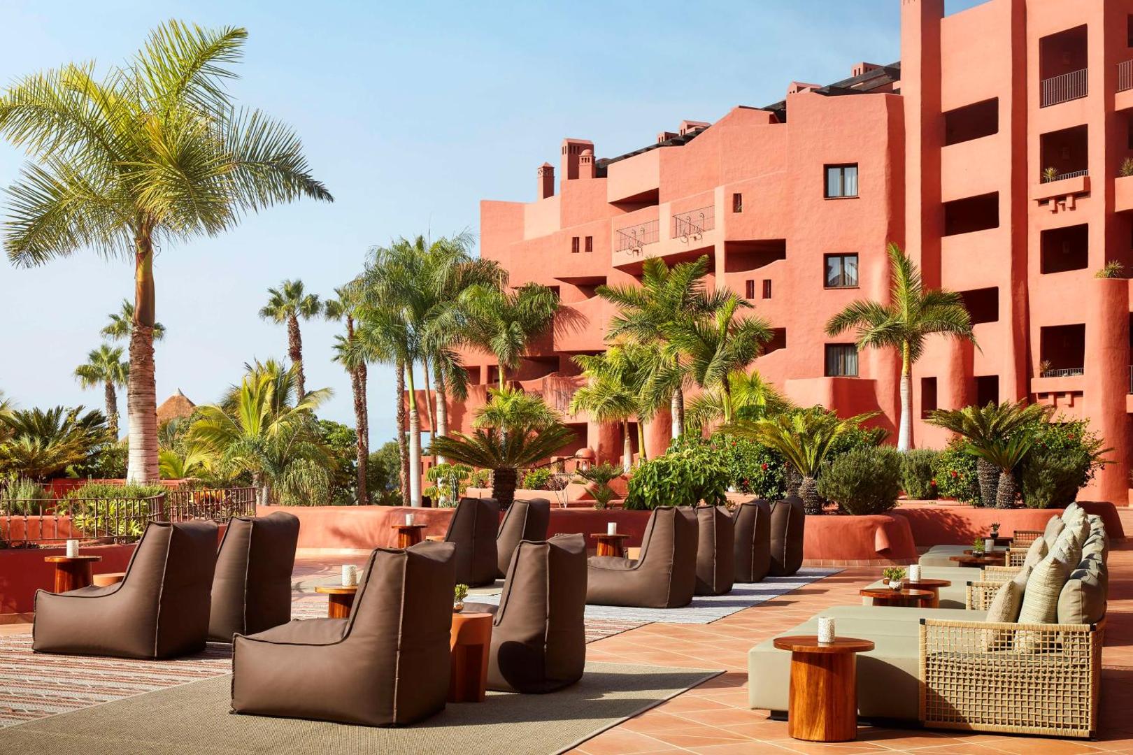 Tivoli La Caleta Tenerife Resort terase ar ērtiem sēžammaisiem un zviļņiem pie viesnīcas sarkanās fasādes un palmām