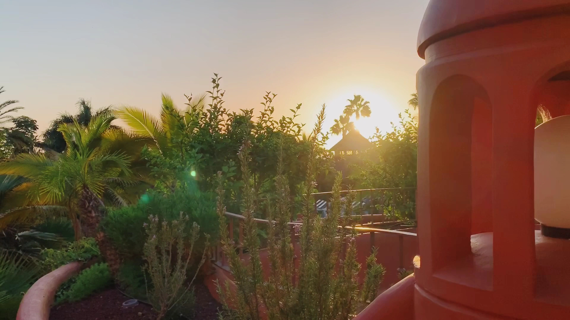 Saulrieta skats no Tivoli La Caleta Tenerife Resort terases caur zaļojošiem augiem un viesnīcas arhitektūras elementiem.