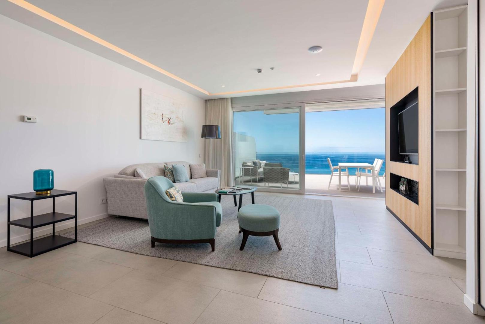 Royal Hideaway Corales Suites viesistaba ar gaišām mēbelēm, dīvānu, atpūtas krēslu un panorāmas logu uz terasi Tenerifē