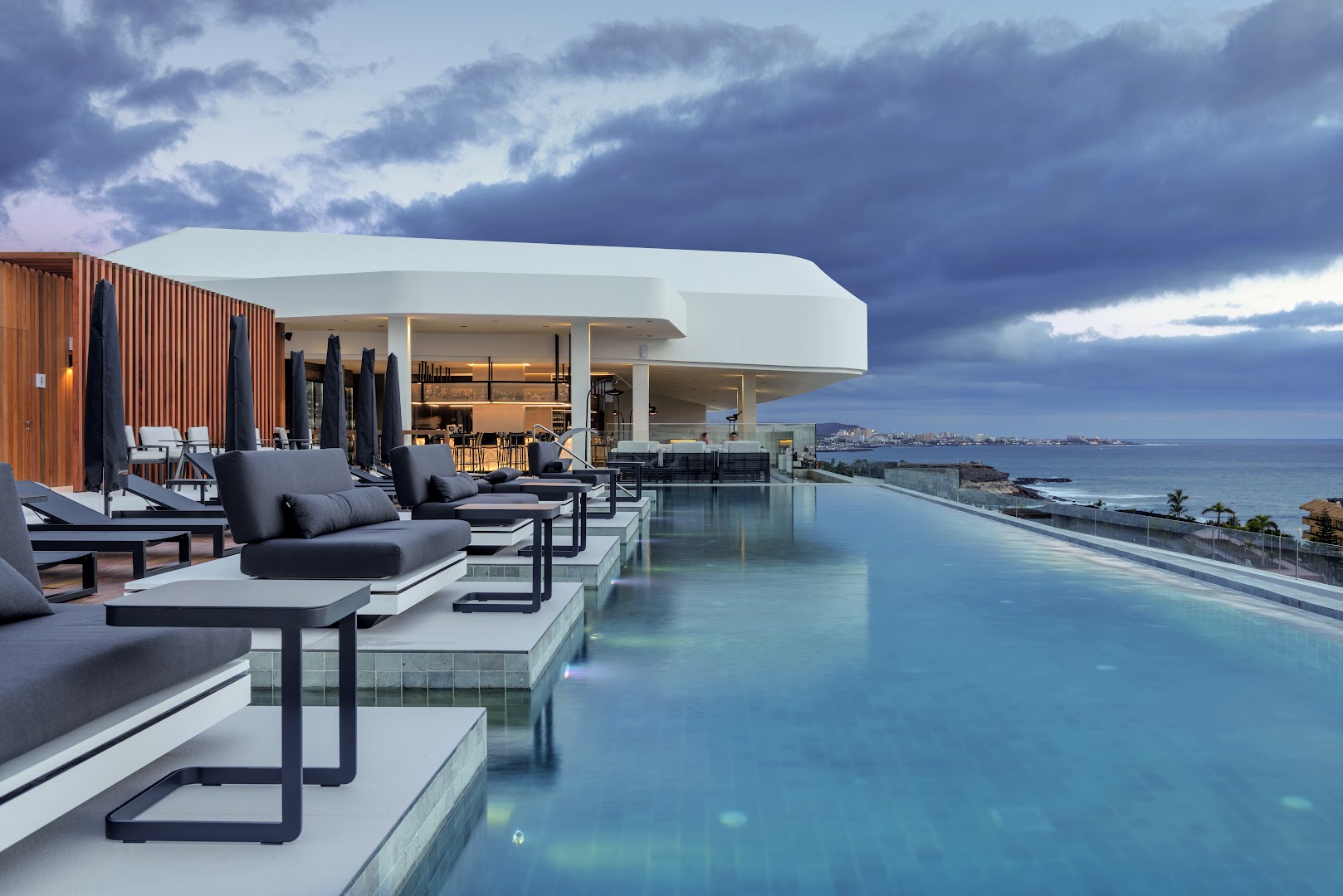 Royal Hideaway Corales Suites bezgalības baseins uz jumta terases ar tumšiem atpūtas krēsliem un skatu uz piekrasti Tenerifē