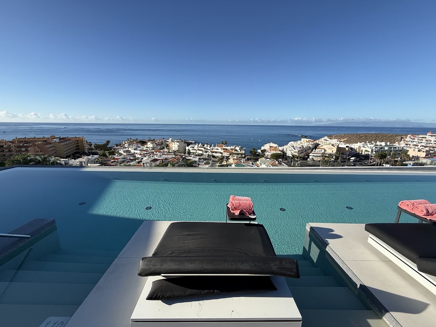 Skats no Royal Hideaway Corales Suites baseina ar pārplūdes malu uz piekrastes pilsētu un okeānu Tenerifē.