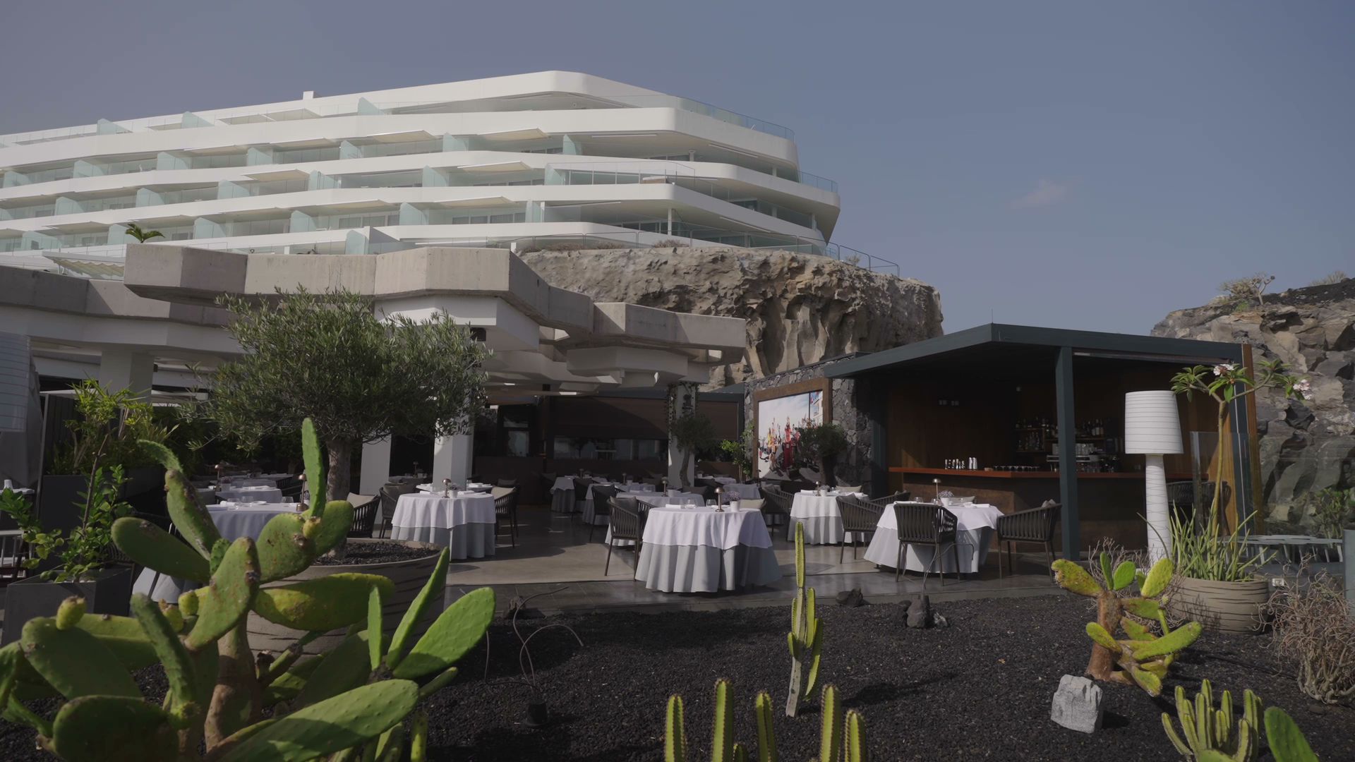 Royal Hideaway Corales Suites viesnīcas modernā arhitektūra un āra restorāns uz klints fona — balti klāti galdi un kaktusu dārzs