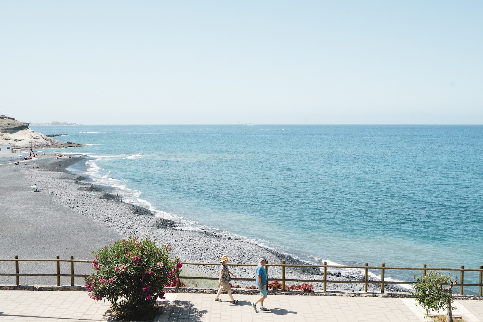 Royal Hideaway Corales Suites skats uz tumšo oļu pludmali un zilo okeānu Tenerifē — pastaigu celiņš piekrastē ar ziedošiem krūmiem