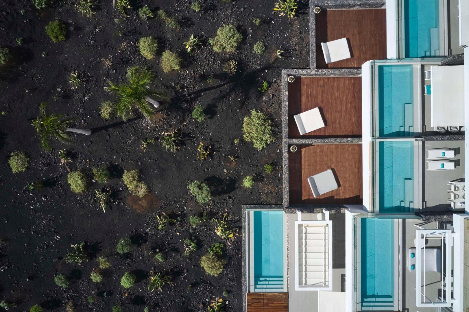Aerofoto uzņēmums ar Royal Hideaway Corales Suites baseiniem un terasēm blakus vulkāniskās augsnes dārzam ar palmām Tenerifē.
