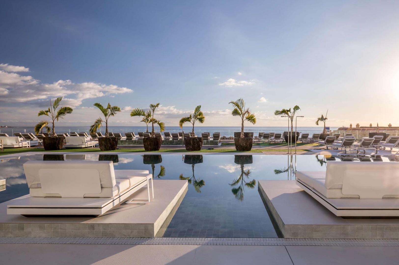 Royal Hideaway Corales Suites āra baseins ar atpūtas krēsliem un palmām saulrietā Tenerifē, skats uz okeānu un horizontu