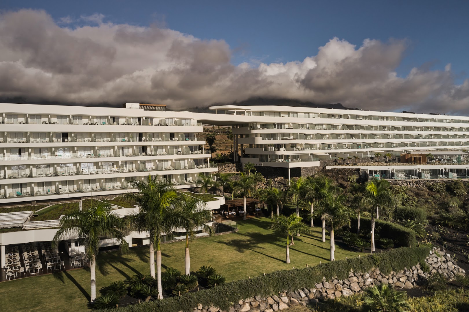 Royal Hideaway Corales Suites viesnīcas ēka un zaļā dārza zona ar palmām Tenerifē, fonā kalni un mākoņi.