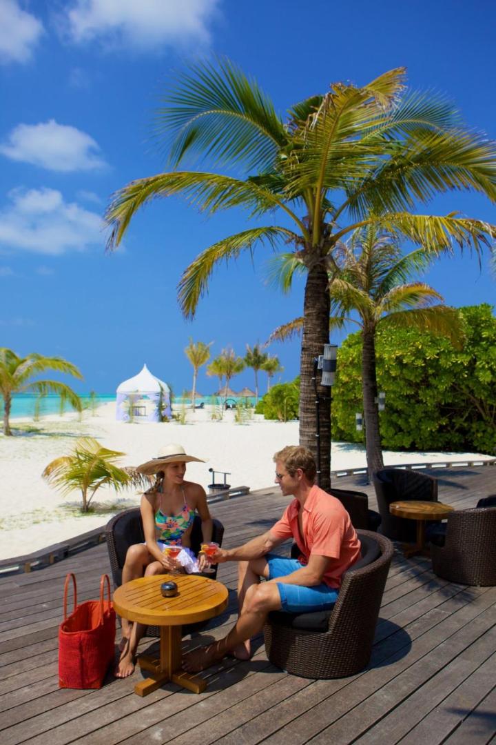 Pāris bauda kokteiļus Kuredu Island Resort & Spa āra terasē pie pludmales — palmas, baltas smiltis un zilas debesis