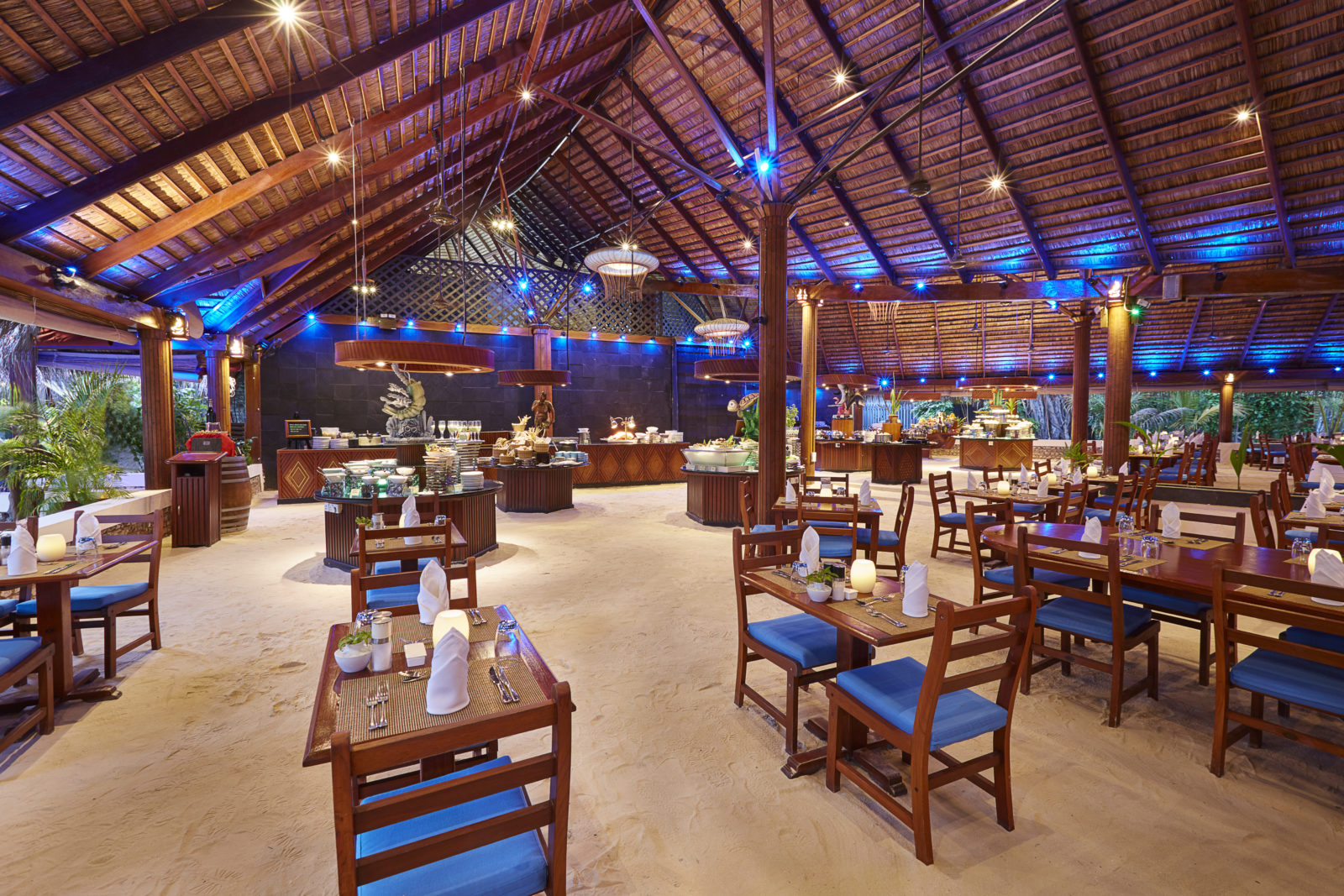 Kuredu Island Resort & Spa restorāns ar smilšu grīdu un bagātīgu bufetes galdu Maldīvu salās — koka interjers un zili krēsli