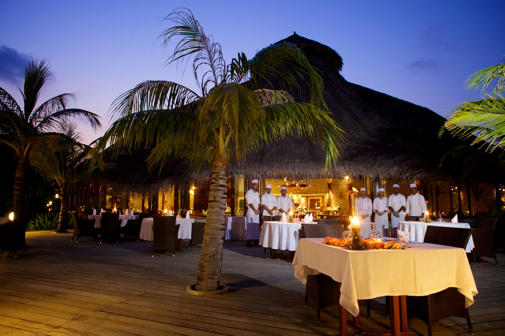 Kuredu Island Resort & Spa restorāna terase vakarā ar personālu un klātiem galdiem zem palmas — tradicionāls niedru jumts