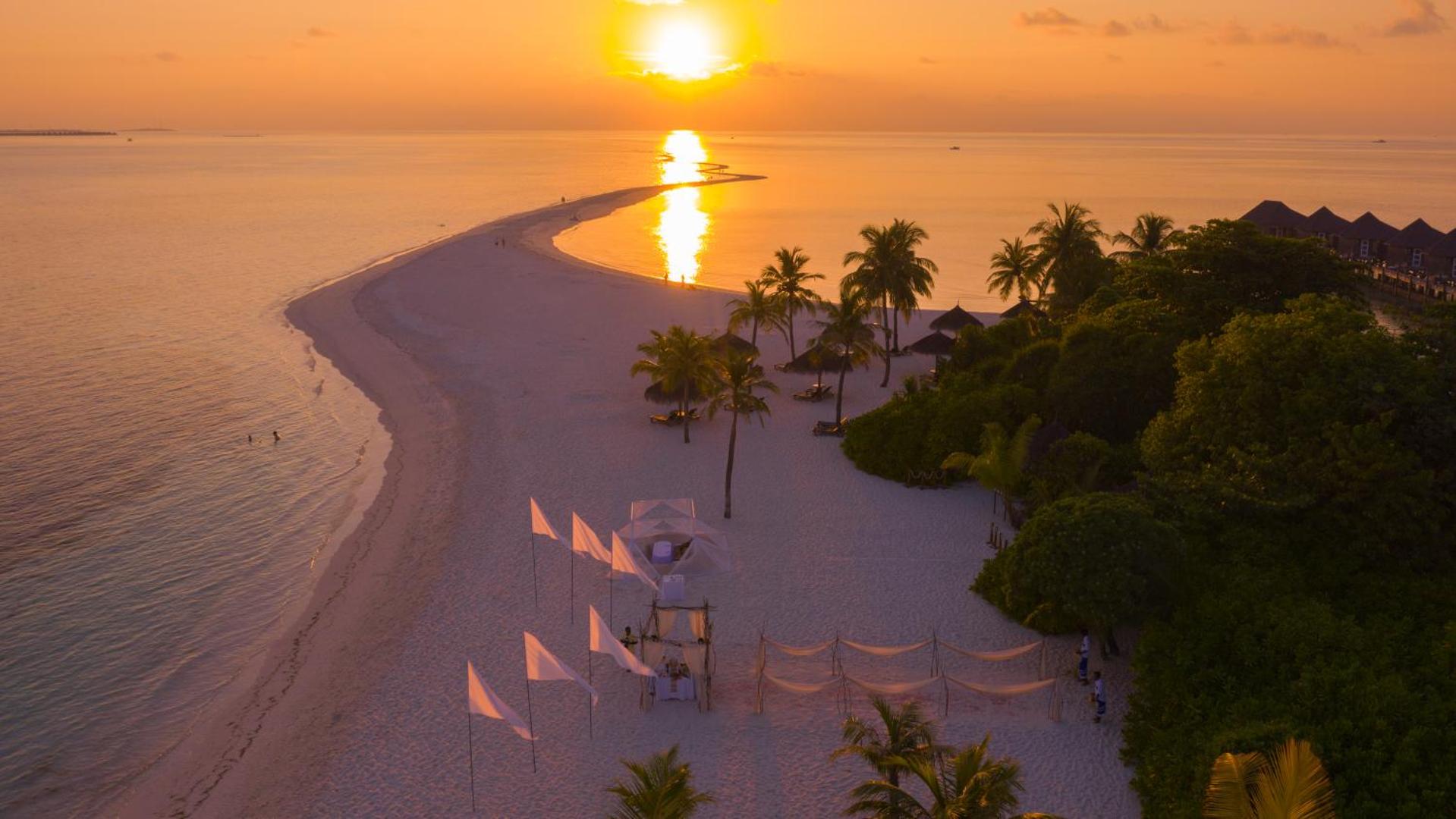 Saulriets Kuredu Island Resort & Spa pludmalē ar baltiem karogiem, palmām un saules atspulgu mierīgajā okeāna ūdenī