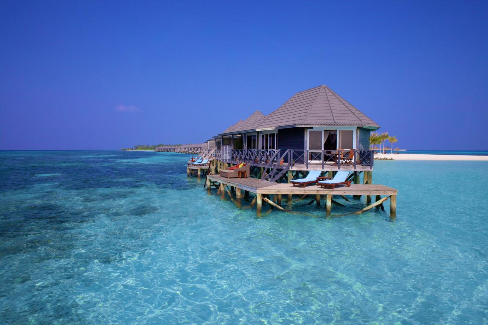 Kuredu Island Resort & Spa ūdens bungalo Maldivu salās virs tirkīzzila okeāna ūdens ar privātu koka terasi un sauļošanās krēsliem