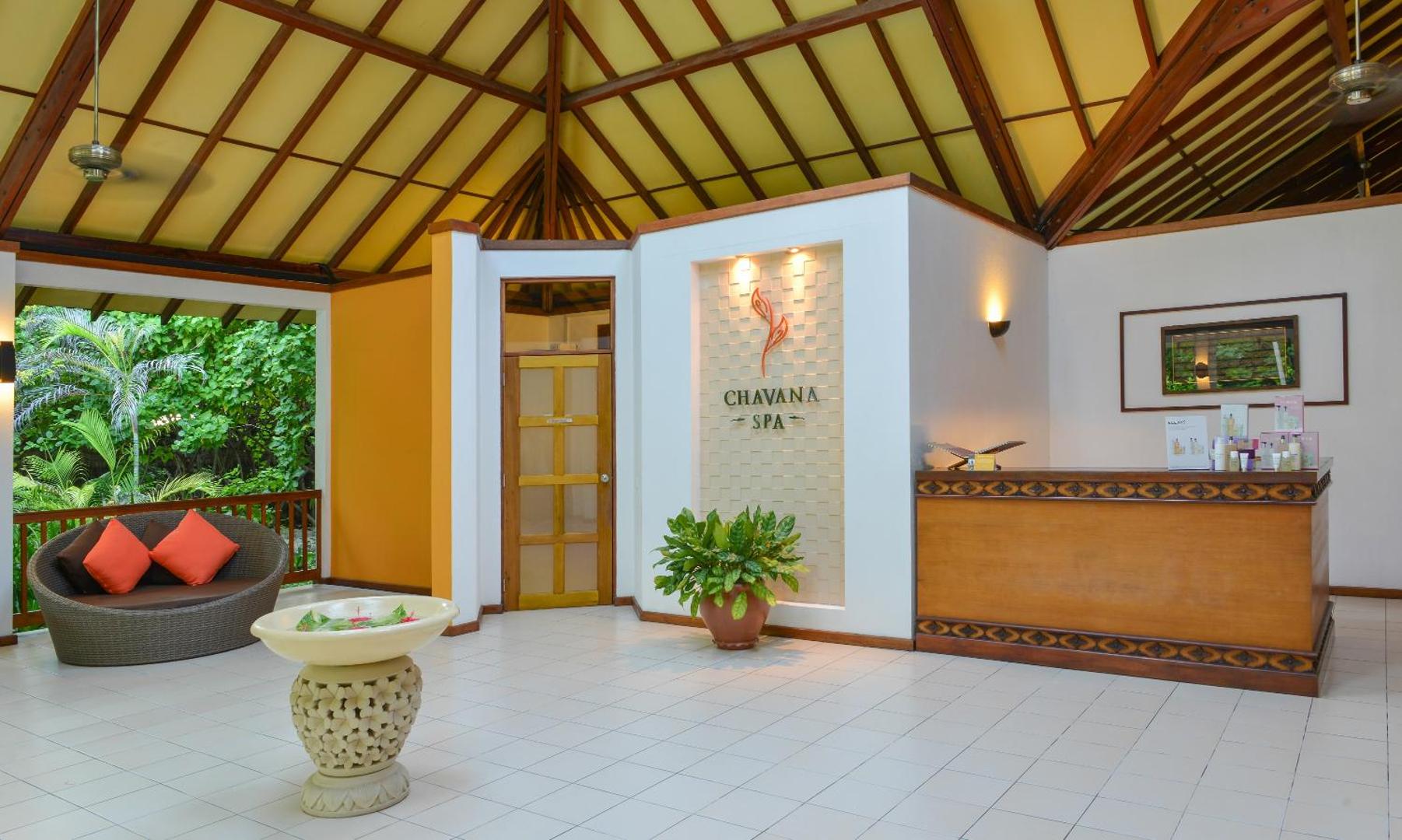 Adaaran Select Meedhupparu Chavana Spa recepcijas zona ar koka apdari, atpūtas dīvānu un tropu augiem Maldīvu kūrortā.