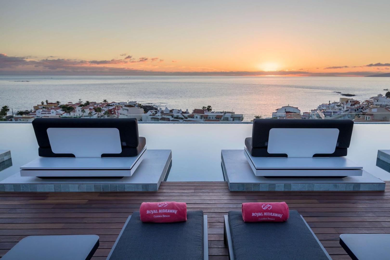 Royal Hideaway Corales Beach terase ar skatu uz okeānu saulrietā Tenerifē — atpūtas krēsli pie baseina ar pārplūdes malu