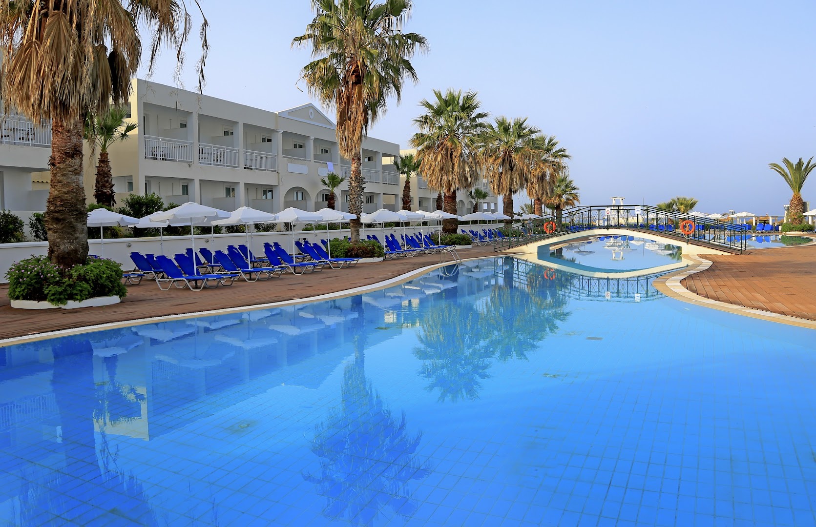 Sandy Beach Resort lielais āra baseins ar tiltiņu, ziliem sauļošanās krēsliem un palmām pie viesnīcas korpusa dienas laikā