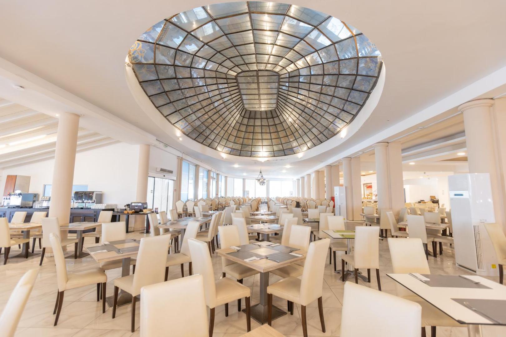 Sandy Beach Resort galvenais restorāns ar iespaidīgu stikla kupolu, gaišām mēbelēm un plašu telpu kopīgām maltītēm Korfu salā.
