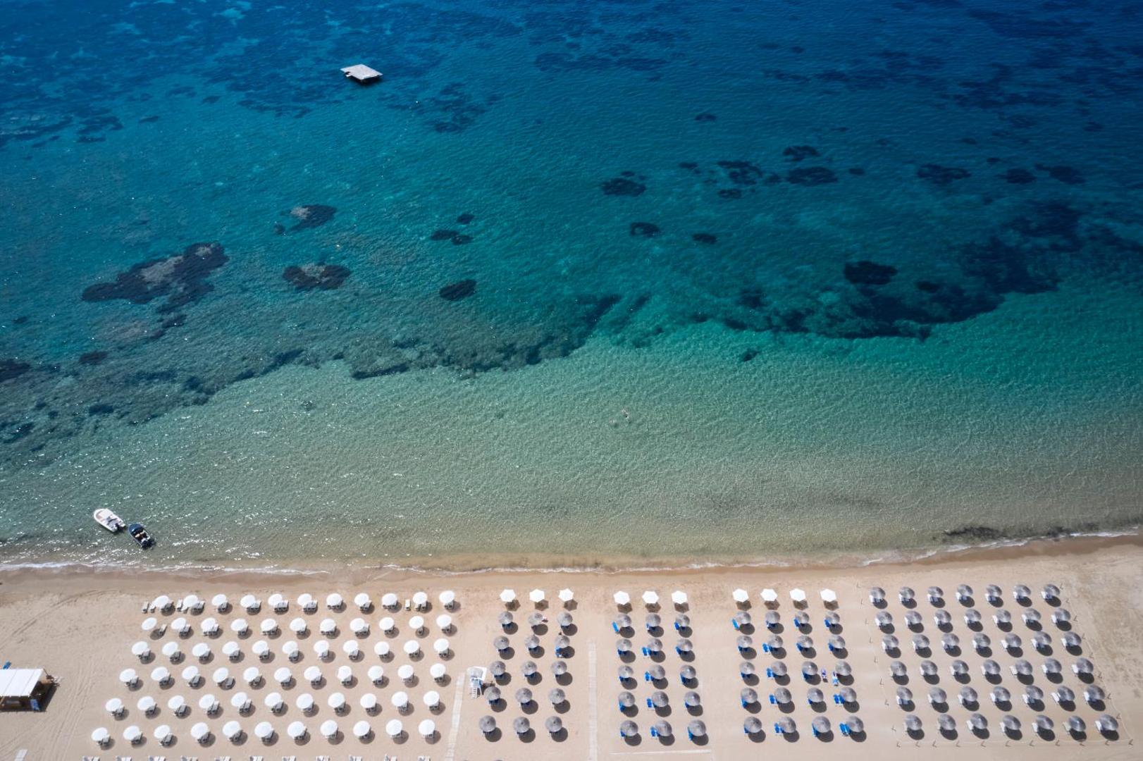 Sandy Beach Resort panorāmas skats no putna lidojuma uz plašo smilšu pludmali, dzidro tirkīzzilo ūdeni un sakārtotām saulessargu rindām.
