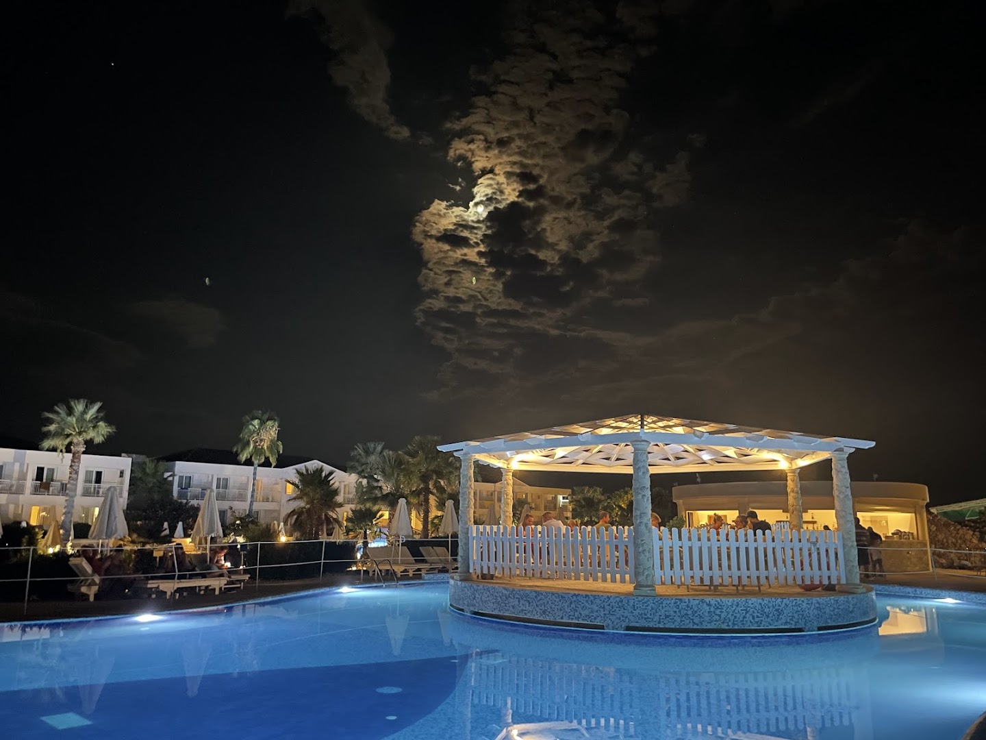 Sandy Beach Resort āra baseins naktī ar izgaismotu lapeni un pilnmēnesi pie debesīm — romantiska atmosfēra kūrorta teritorijā