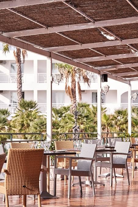 Sandy Beach Resort mājīga āra terase ar pītiem krēsliem zem dabīga materiāla nojumes, skats uz palmu dārzu un viesnīcas korpusu.
