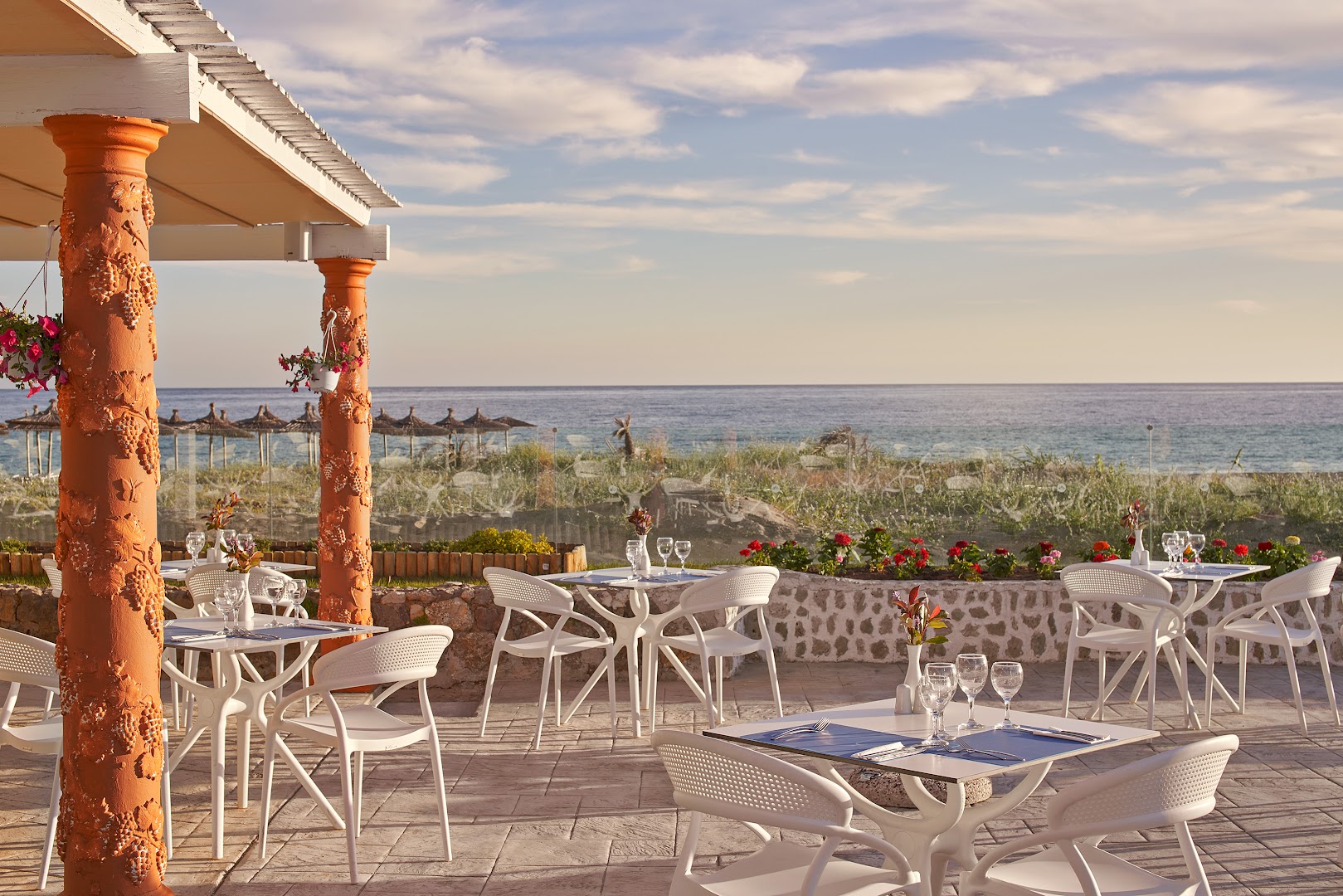 Sandy Beach Resort āra terases restorāns ar skatu uz jūru — balti galdi un krēsli, dekoratīvas kolonnas un saulrieta gaisma