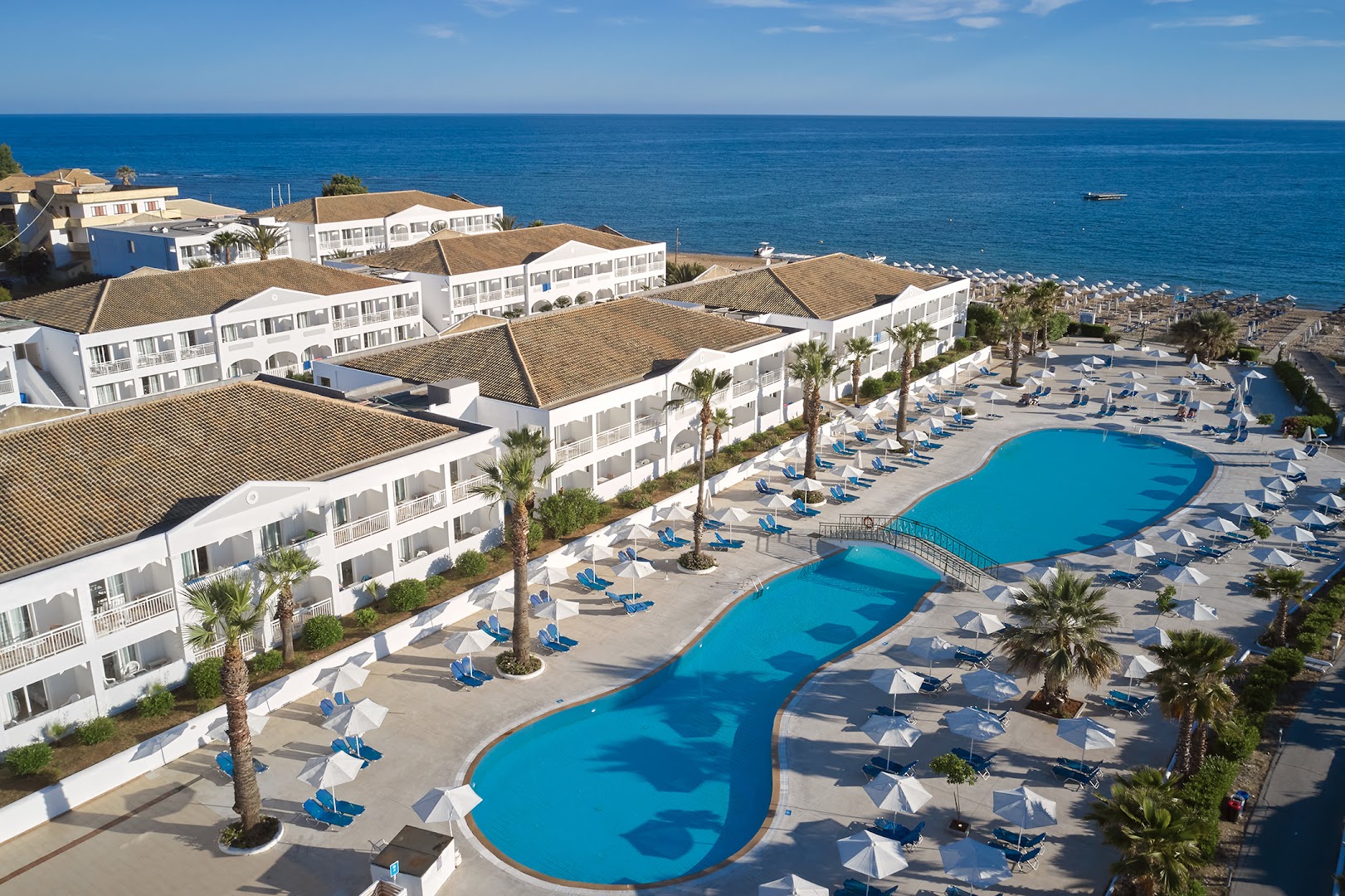 Sandy Beach Resort centrālais baseins ar saulessargiem un palmām Korfu — fonā redzamas viesnīcas korpusa ēkas ar balkoniem