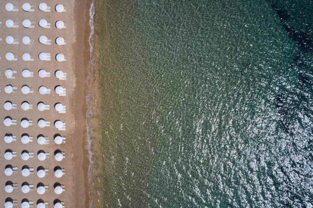 Sandy Beach Resort pludmales tuvplāns no augšas, parādot smilšu joslu, baltus saulessargus un saules atspulgus dzidrajā jūras ūdenī.