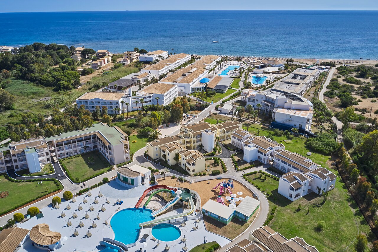 Sandy Beach Resort panorāmas skats no putna lidojuma Korfu salā — plaša teritorija ar baseiniem, bērnu akvaparku un tiešu pieeju jūrai