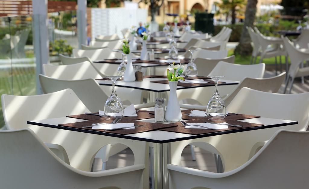 Sandy Beach Resort āra terases restorāns ar baltiem krēsliem un servētiem galdiem, ko ieskauj zaļojošs dārzs un saules gaisma.