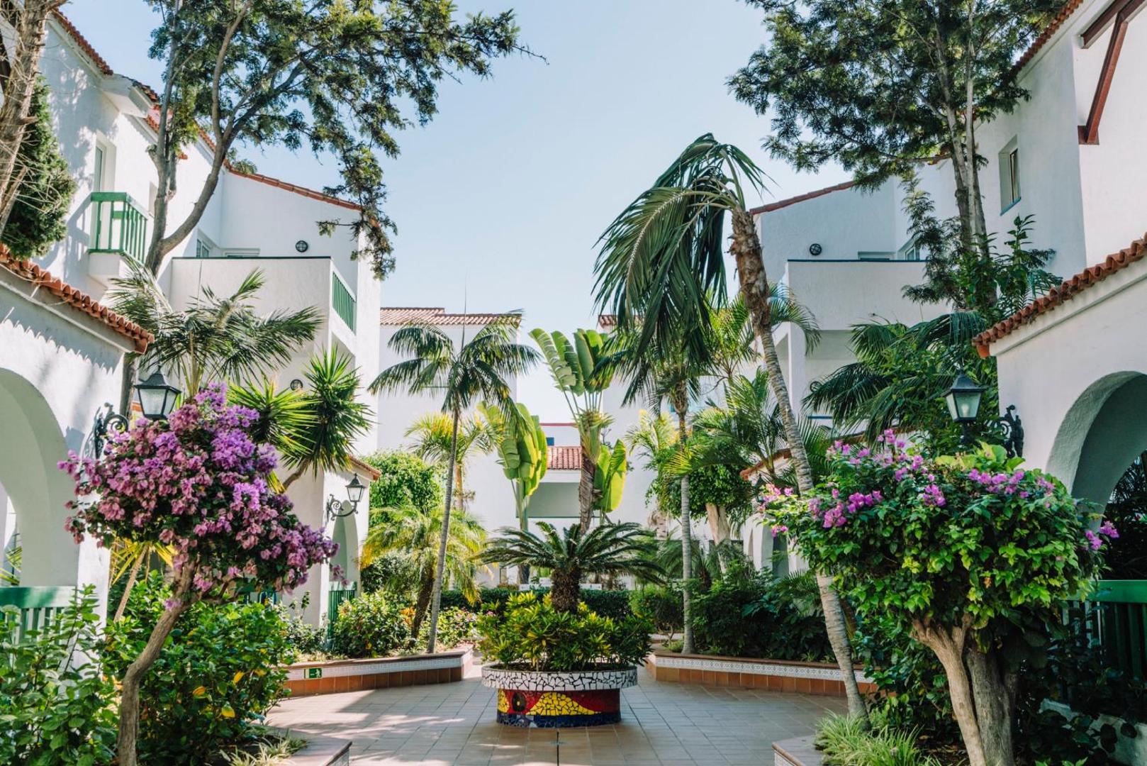 Viesnīcas Park Club Europe iekšpagalms Tenerifē ar baltām ēkām, palmām un ziedošiem bugenvilleju krūmiem saulainā dienā.