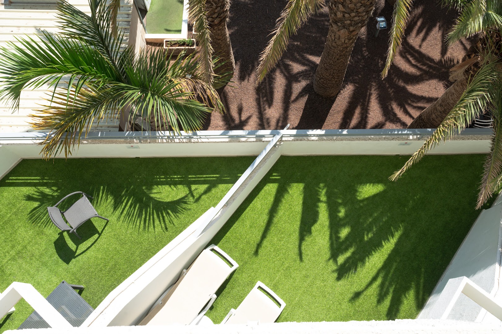 Skats no augšas uz Ole Tropical Tenerife numura terasi ar mākslīgo zālāju, krēslu un palmu ēnu rakstiem