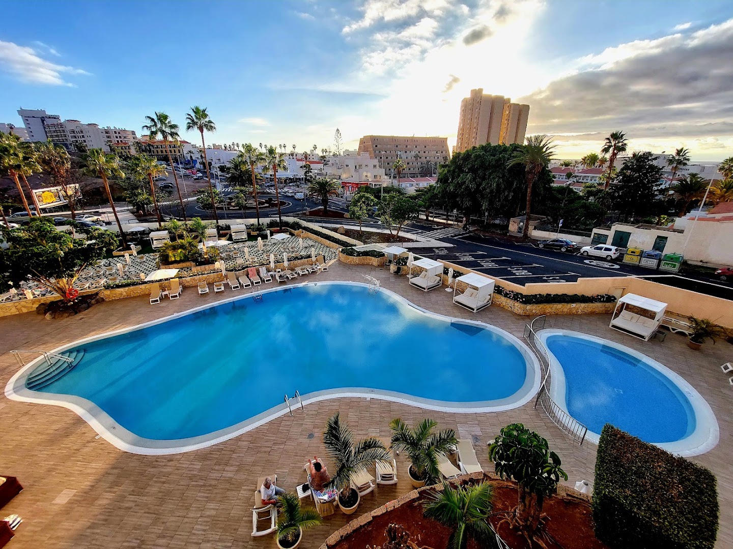 Skats no augšas uz Ole Tropical Tenerife baseinu zonu saulrietā ar pilsētas panorāmu un palmām fonā