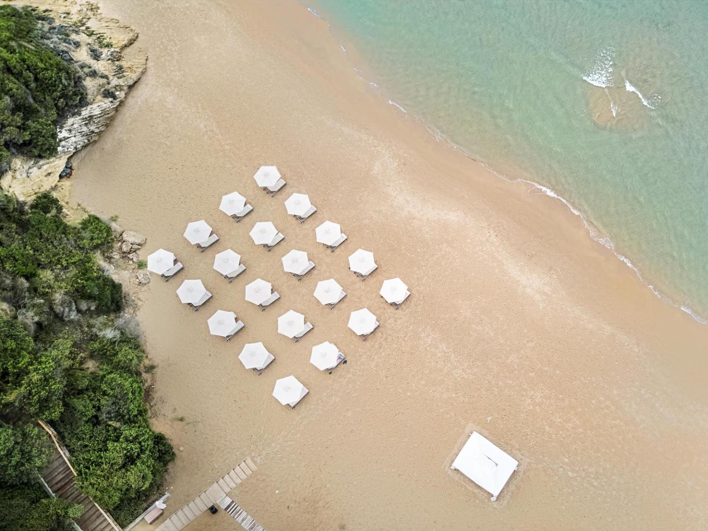 Grecotel Mandola Rosa pludmales skats no augšas ar simetriski izvietotiem baltiem saulessargiem un dzidru jūras ūdeni.