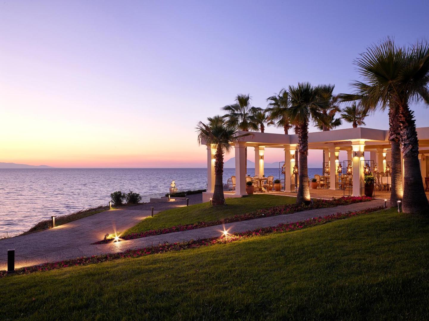 Grecotel Mandola Rosa ēdienkartes restorāna terase saulrieta laikā ar skatu uz Jonijas jūru, palmām un romantisku apgaismojumu.