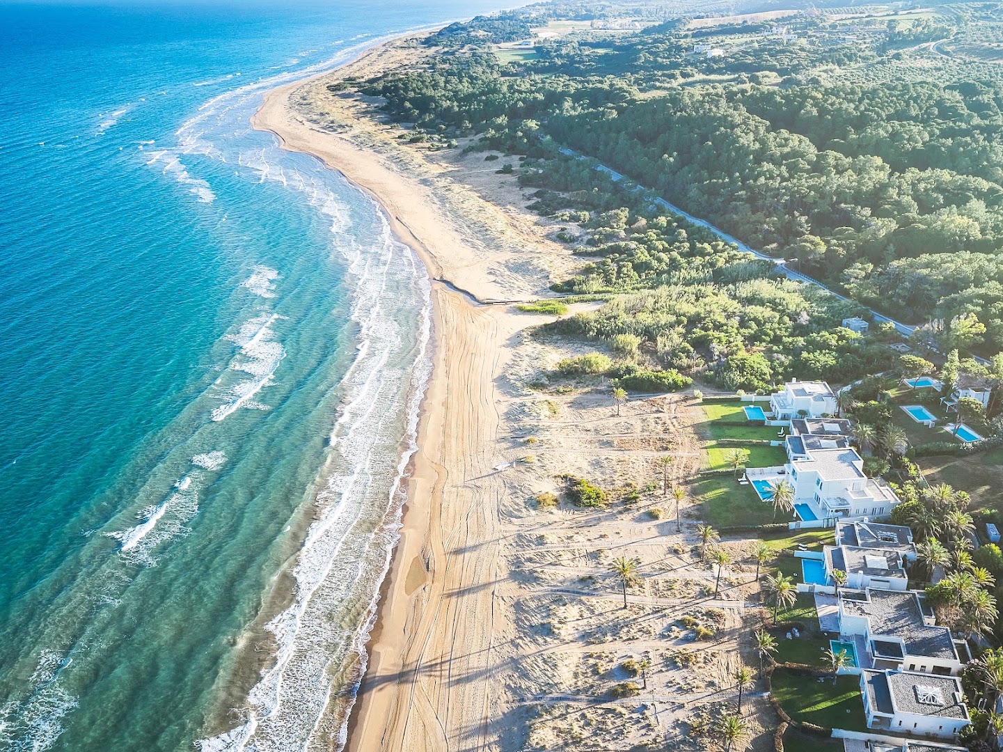 Grecotel Mandola Rosa aerofotoattēls ar plašu smilšu pludmali un luksusa villām Peloponēsas piekrastē, Grieķijā, ko ieskauj zaļš priežu mežs.