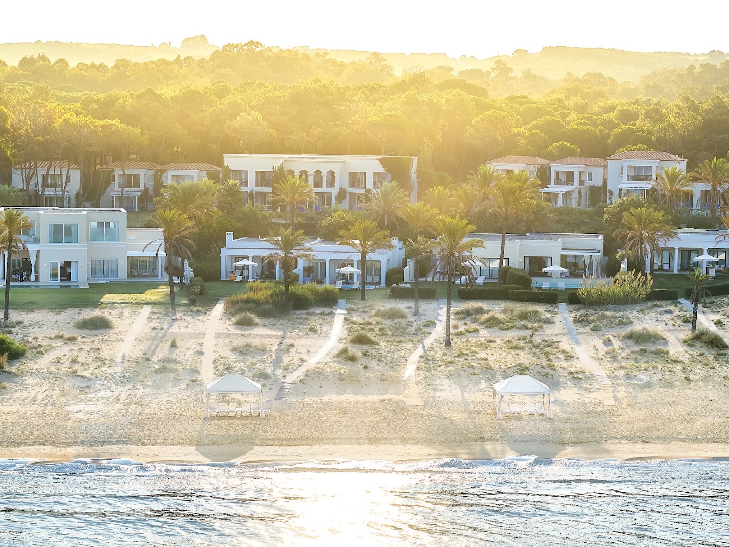 Grecotel Mandola Rosa kūrorta skats no jūras puses saullēktā, parādot villas, kāpas un priežu mežu fonā.