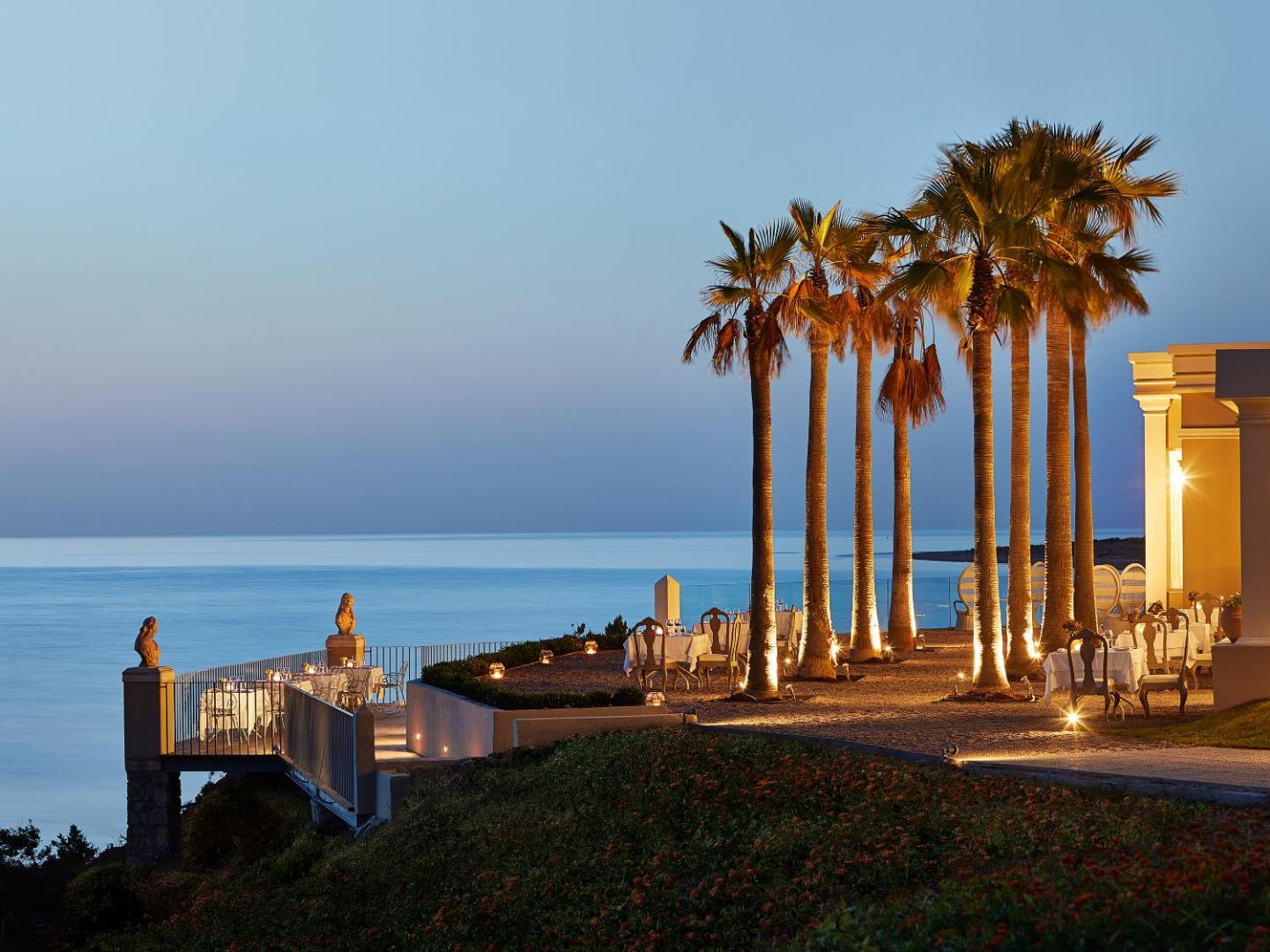 Grecotel Mandola Rosa restorāna terase vakarā ar palmām un skatu uz Jonijas jūru — eleganti klāti galdi un silts apgaismojums.