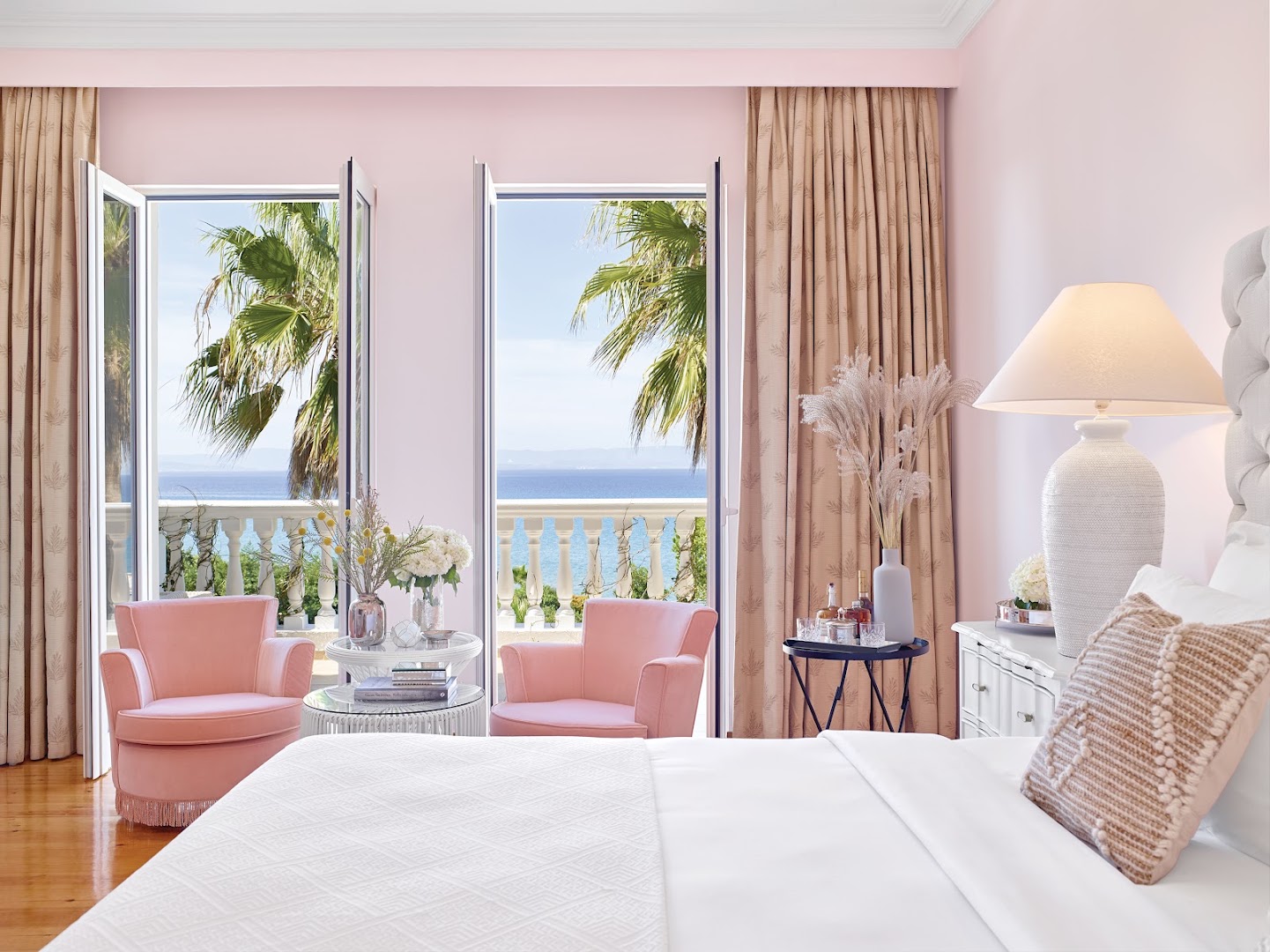 Grecotel Mandola Rosa numurs ar rozā sienām un skatu uz jūru — divi rozā atpūtas krēsli pie balkona durvīm un palmas