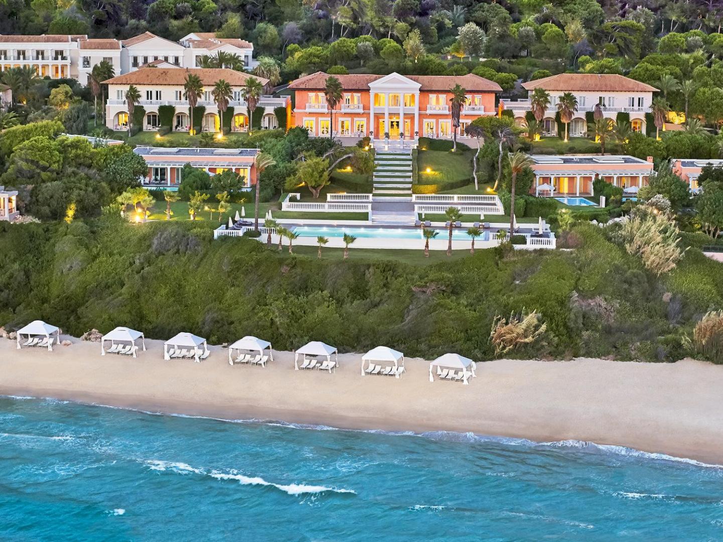 Grecotel Mandola Rosa galvenā ēka klasiskā arhitektūras stilā uz pakalna ar skatu uz baseinu un pludmali vakarā.