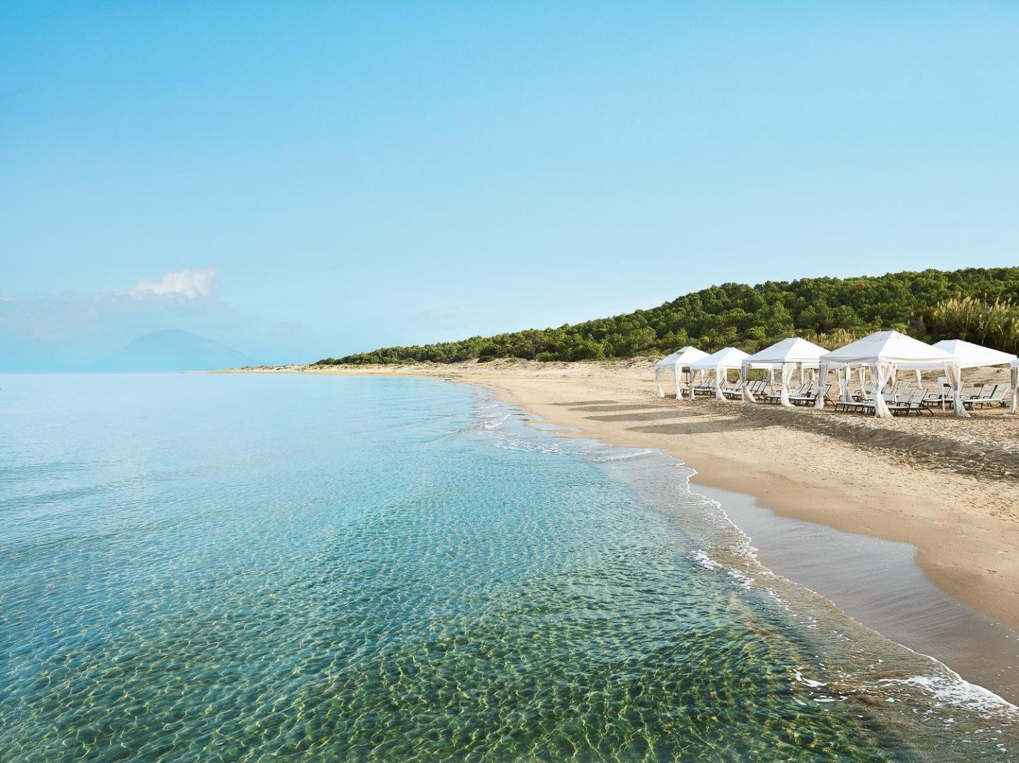 Grecotel Mandola Rosa pludmale ar baltām teltīm un sauļošanās krēsliem pie dzidra, sekla ūdens saulainā dienā.