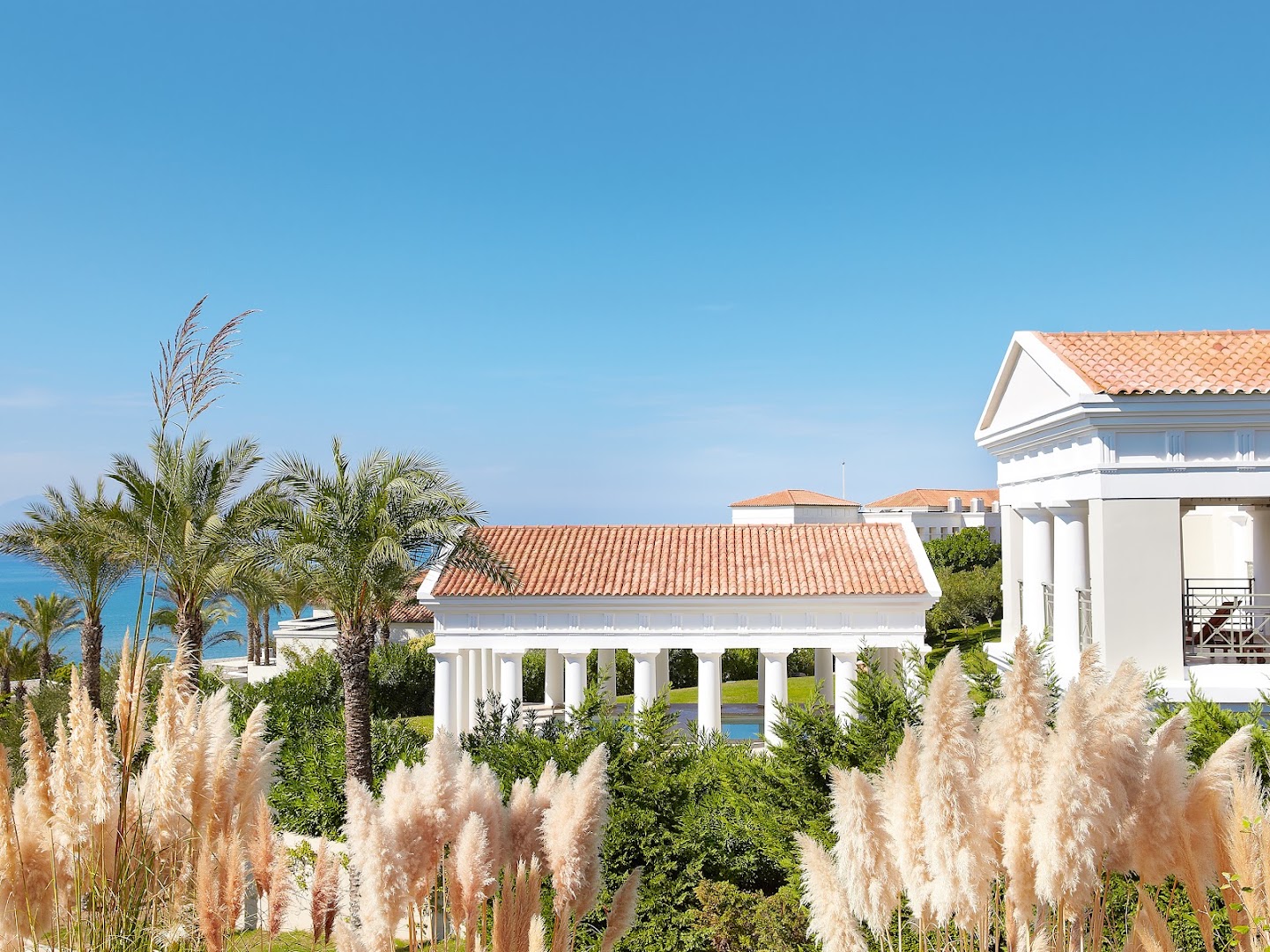 Grecotel La Riviera klasiskā arhitektūra ar baltām kolonnām un sarkaniem dakstiņu jumtiem starp palmām un dekoratīvajiem augiem