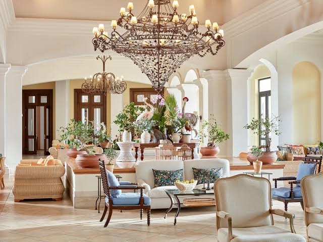 Grecotel La Riviera viesnīcas vestibils Peloponēsā ar greznām lustrām, klasiskām mēbelēm un dekoratīviem augiem gaišā un plašā interjerā.